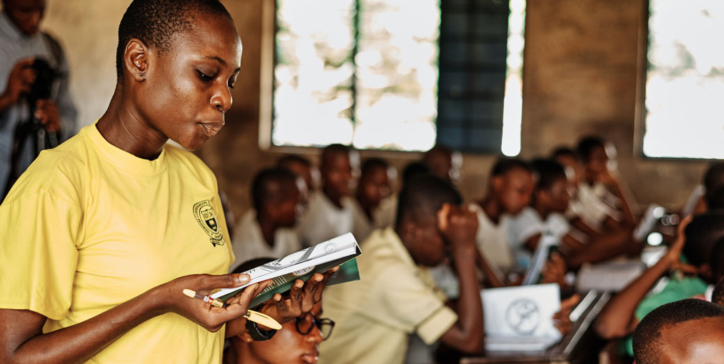 A teacher in a school with textbook and students in the background